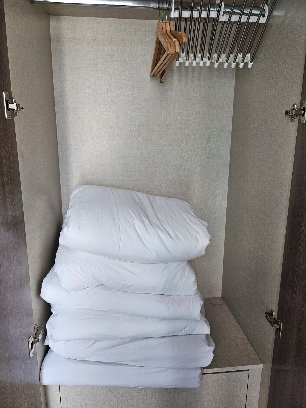 A neatly stacked set of white pillows in a corner of a room with a neutral-toned wall and a metal rack with a brown item hanging above.