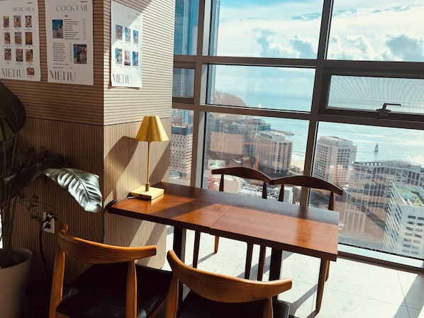 Habitación de hotel en un edificio alto, con una mesa y sillas de madera, una lámpara y vistas a la ciudad y al océano.