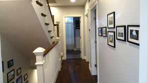 A hallway with a wooden staircase, white walls, and framed pictures. - Park-Side Home in Jamaica Plain (Boston)