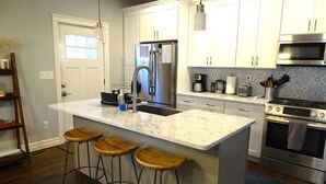 A modern kitchen with white cabinets, a stainless steel refrigerator, a Bosch oven, and a marble countertop. There are three wooden stools at the island. - Park-Side Home in Jamaica Plain (Boston)