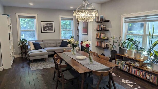 A modern living room with a wooden dining table, a sofa, and a bookshelf filled with books. - Park-Side Home in Jamaica Plain (Boston)