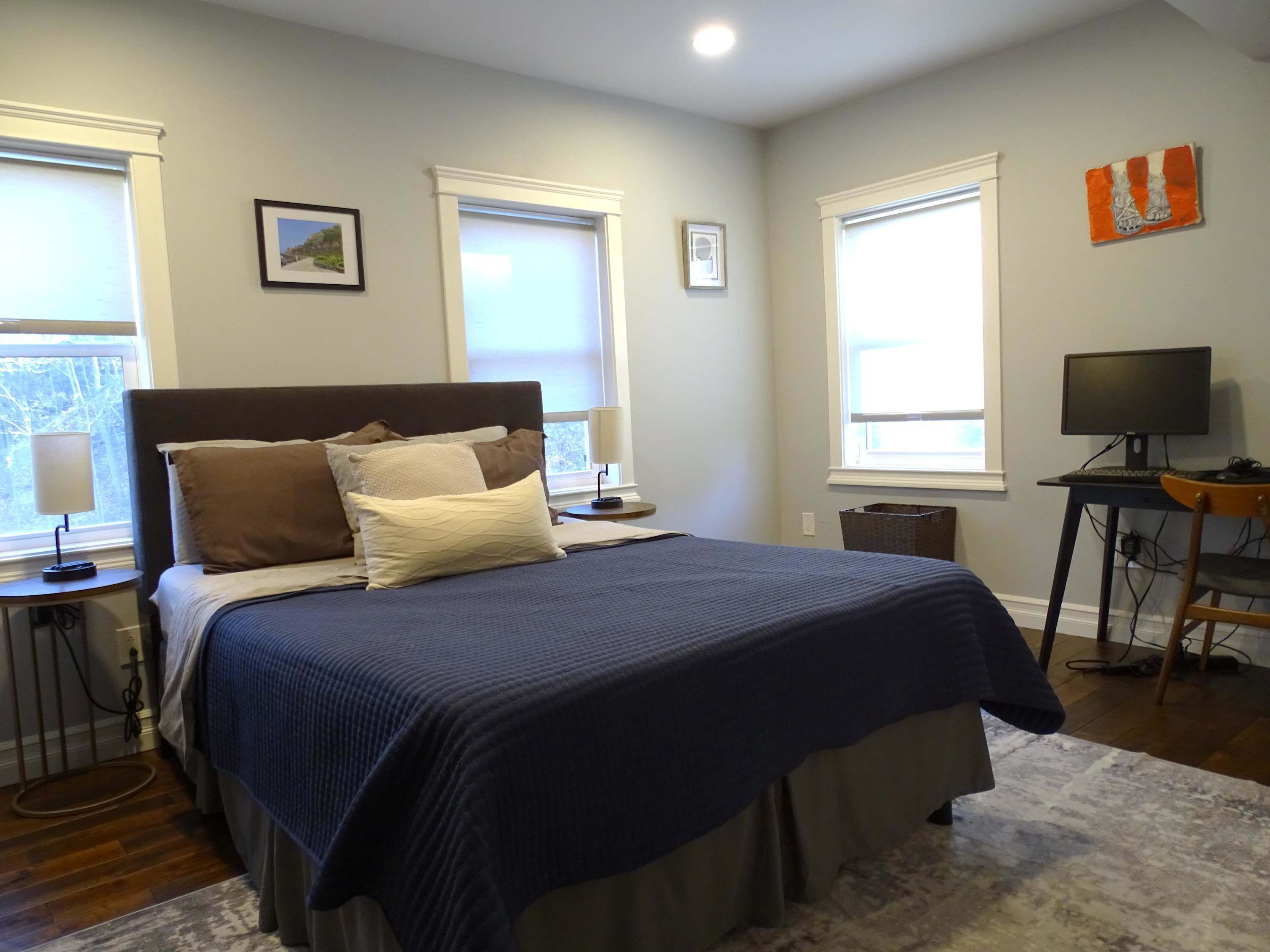 A bedroom with a large bed, two bedside tables, a desk with a computer, and windows with blinds.