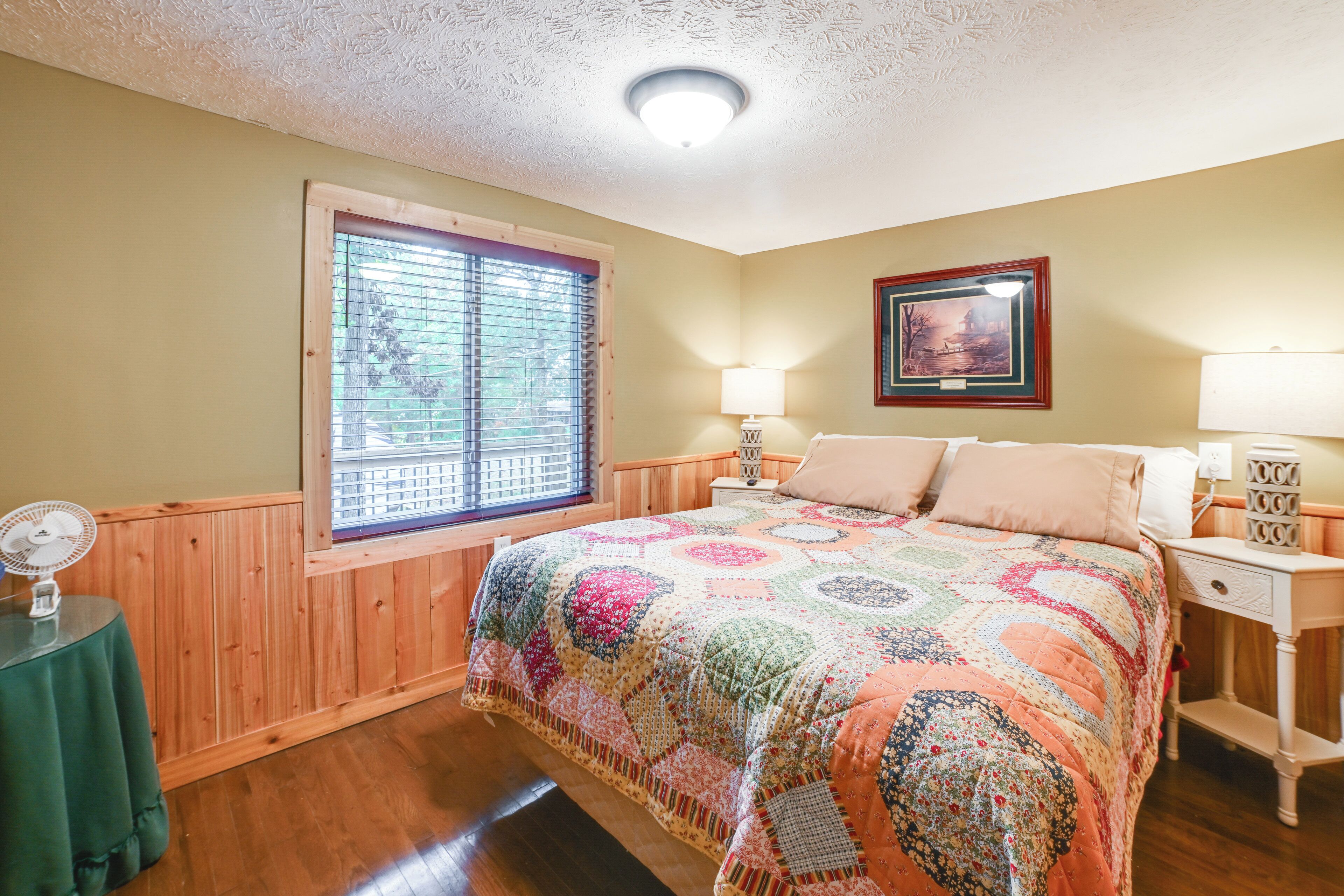 A bedroom with a wooden paneled wall, a large window with blinds, a colorful quilted bedspread, a bedside table with a lamp, and a framed picture on the wall.