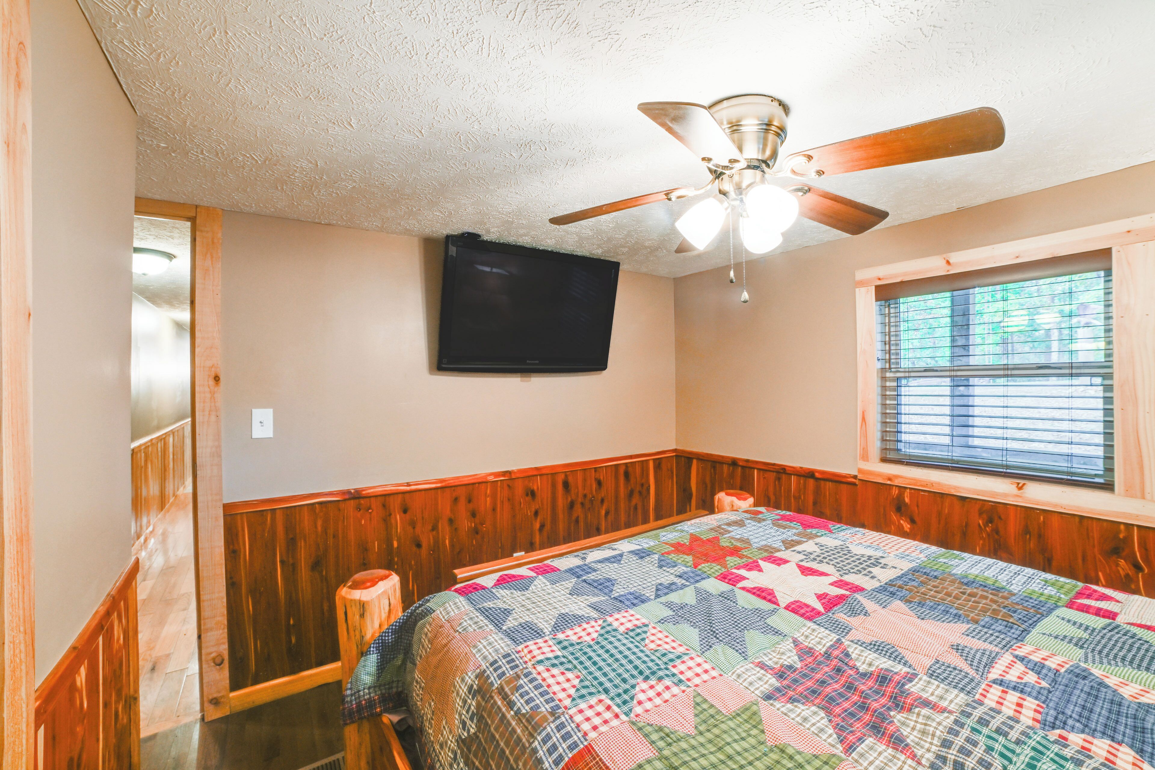 A bedroom with a wooden floor, a bed with a colorful quilt, a ceiling fan, a flat-screen TV, and a window with blinds.
