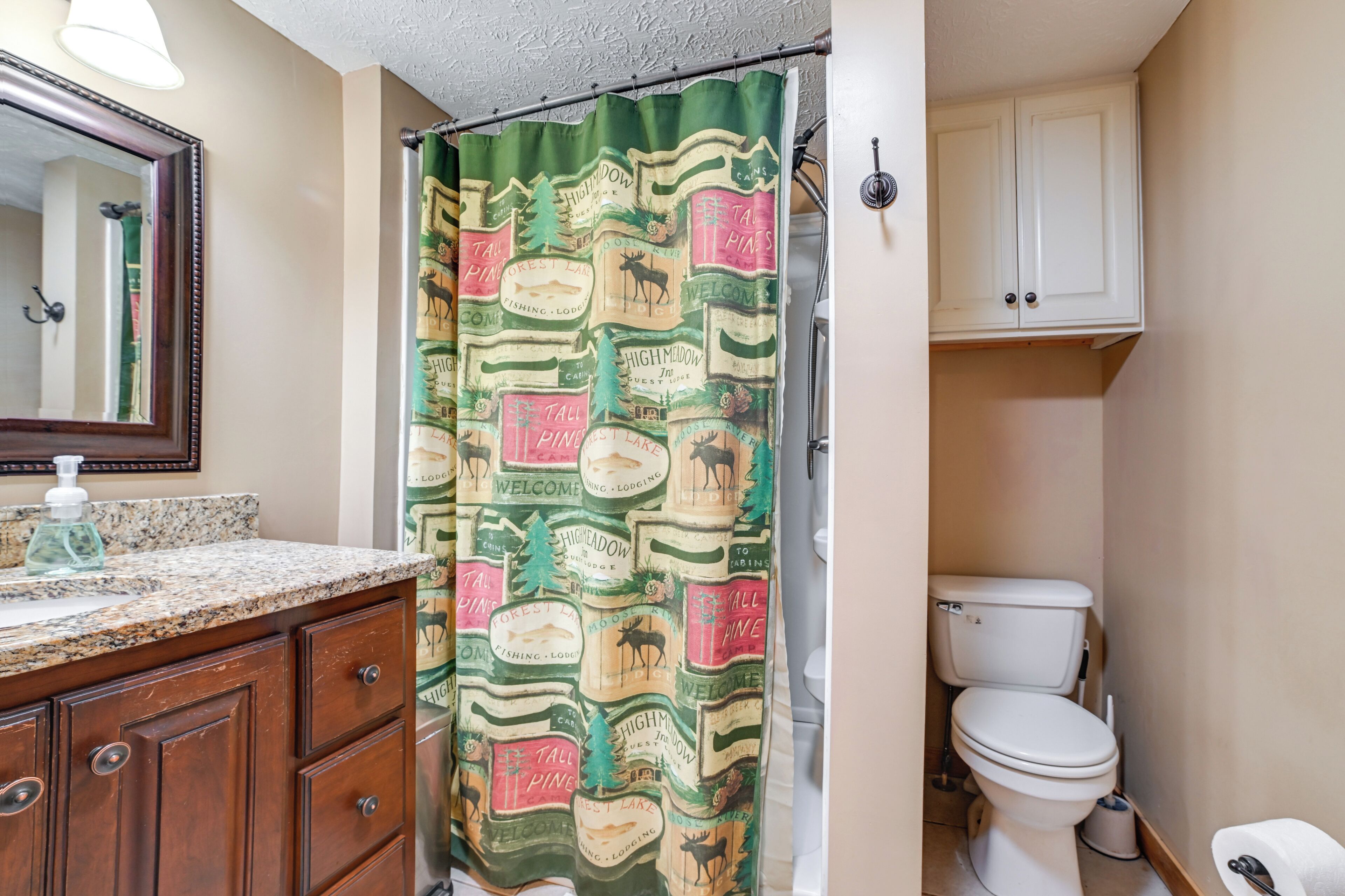 Une salle de bain avec un rideau de douche orné d’un motif représentant une forêt et des animaux sauvages, des toilettes et un meuble-lavabo en bois avec un miroir et un distributeur de savon.