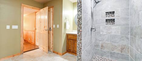 A bathroom with a shower, tiled floor, wooden cabinets, and a door leading to another room.