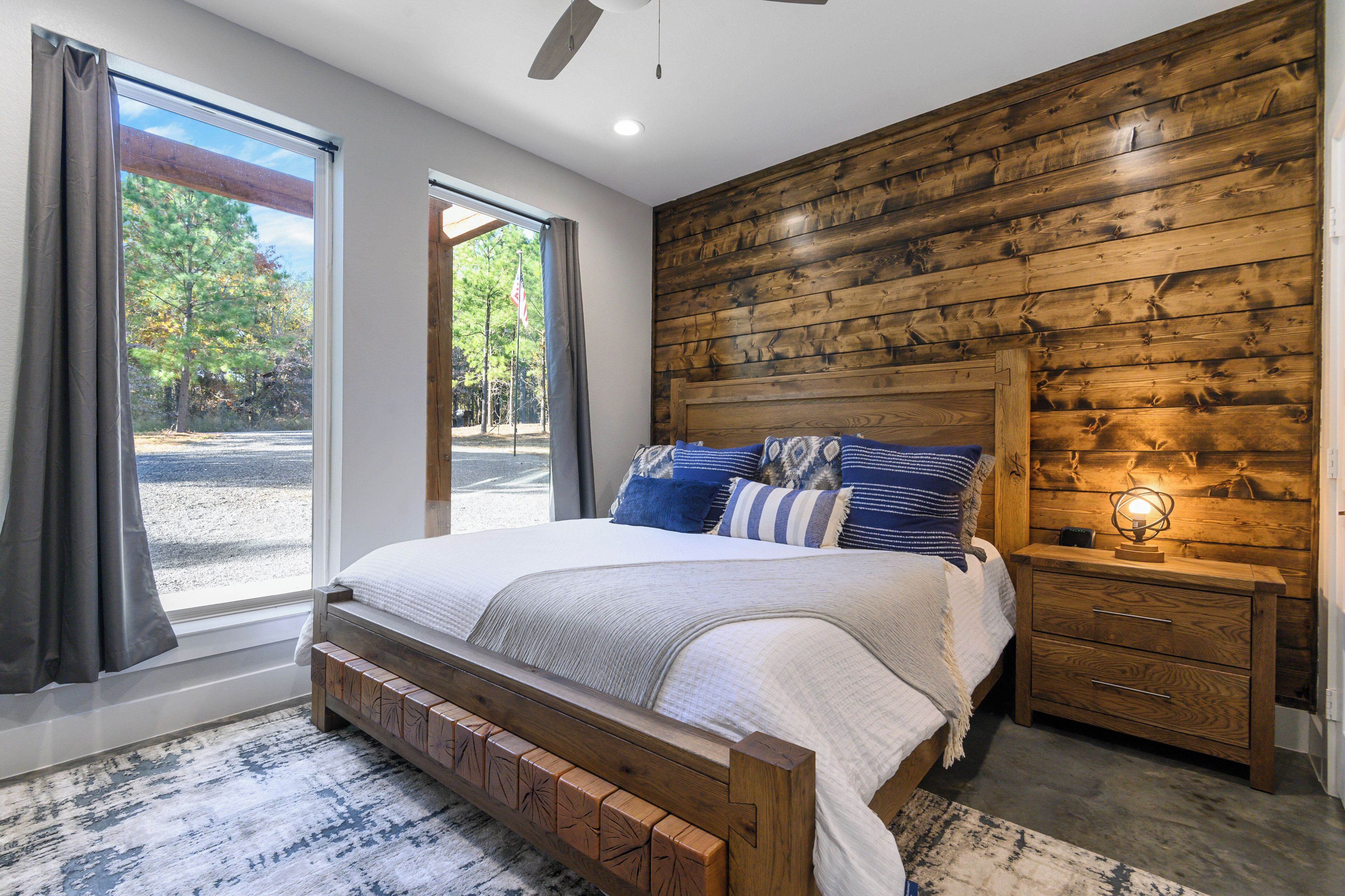 A bedroom with a wooden headboard, a large bed with multiple pillows, a nightstand with a lamp, and a view of trees through a window.