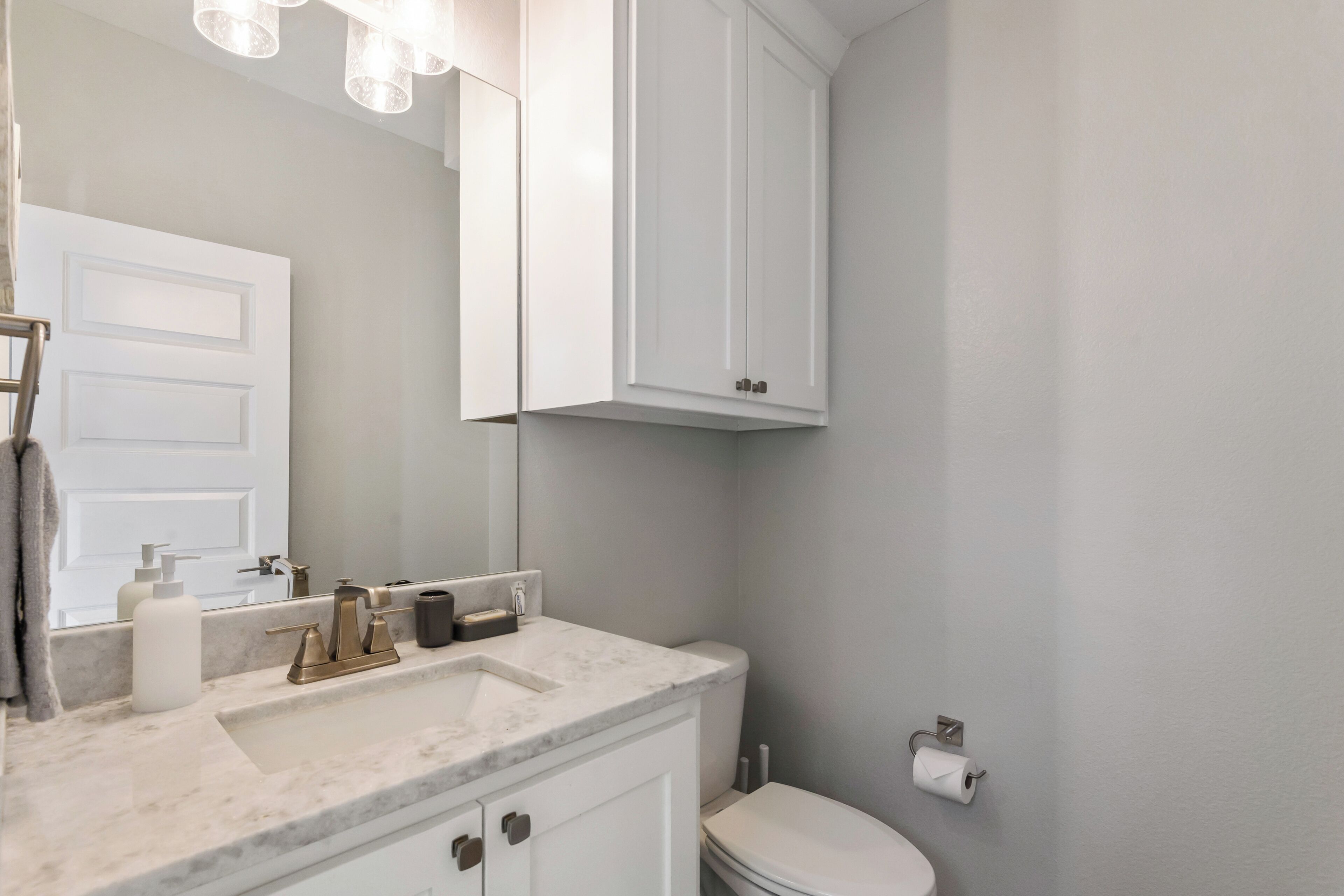 A bathroom with a white sink, mirror, and cabinet, a toilet, and a towel hanging on the door.
