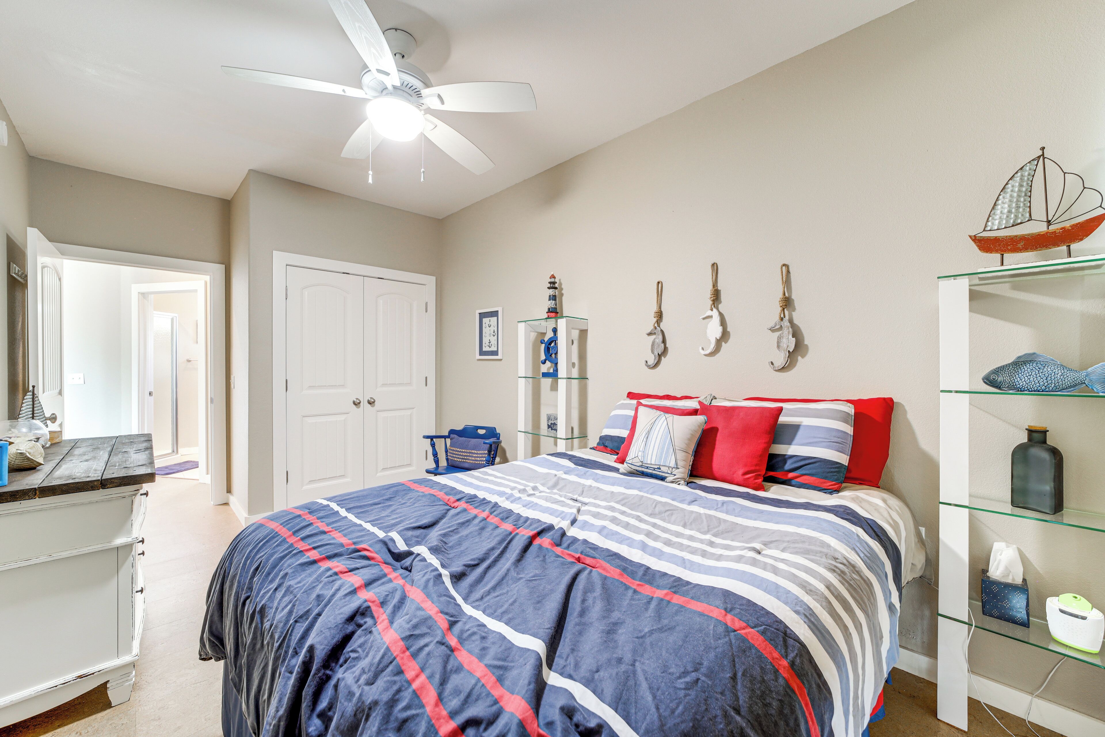 A bedroom with a bed, a ceiling fan, a dresser, a glass shelf with decorative items, and a door leading to another room.