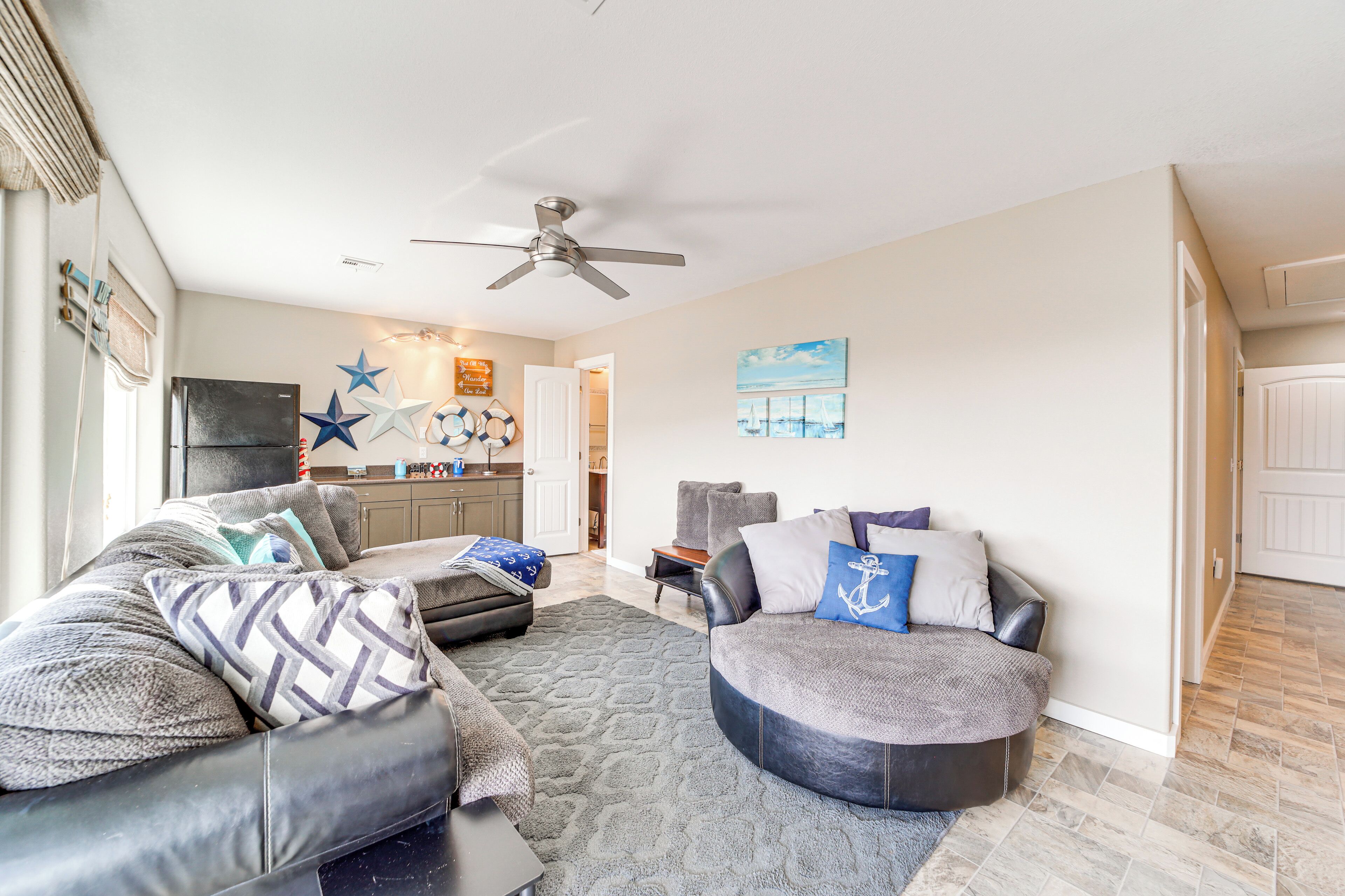 A living room with a sofa, ottoman, and a ceiling fan. There is a kitchen in the background.