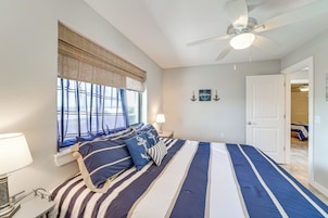 A bedroom with a large bed, a ceiling fan, a window with blinds, a bedside table with a lamp, and a door leading to another room.