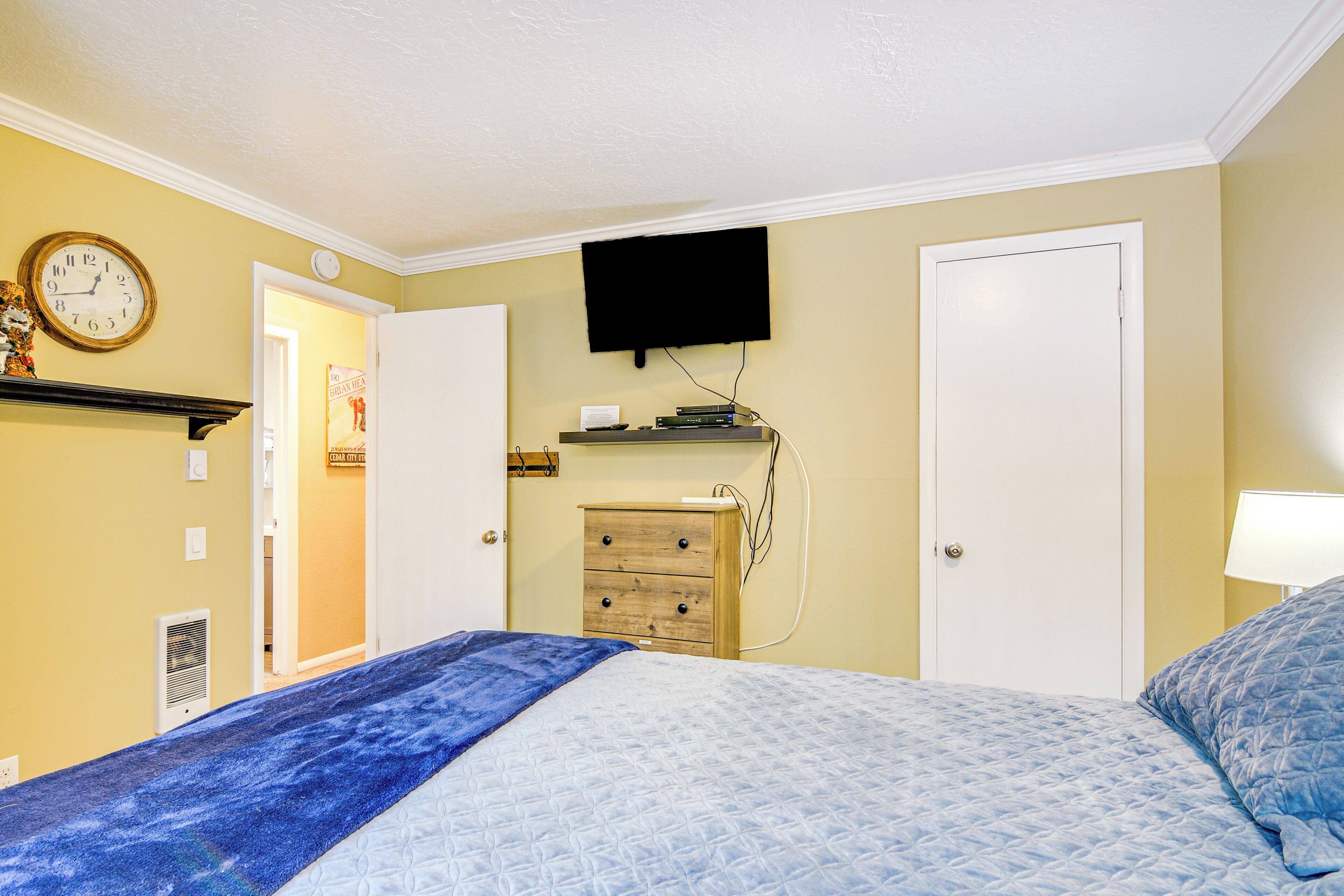 A bedroom with a bed, a dresser, a television mounted on the wall, and a clock on the wall.
