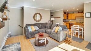 A modern living room with a gray sectional sofa, a round coffee table, and a small wooden side table. A kitchen with wooden cabinets and a white dining table is visible in the background. - Walk to Giant Steps & Hot Tub: Brian Head Condo! (Brian Head)