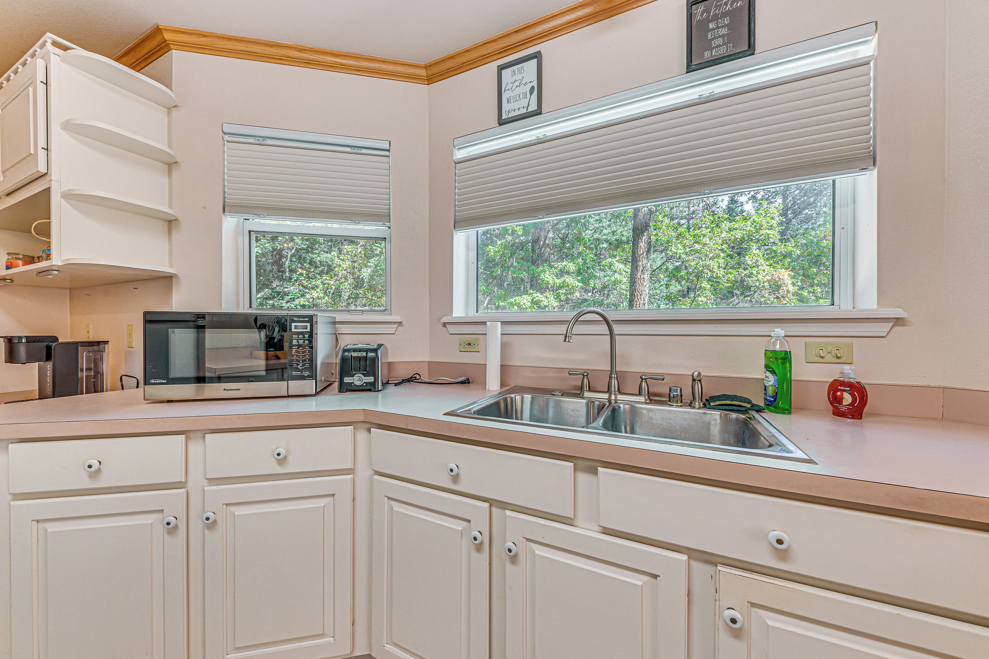 A kitchen with white cabinets, a microwave, a toaster, a sink, and two windows with blinds.