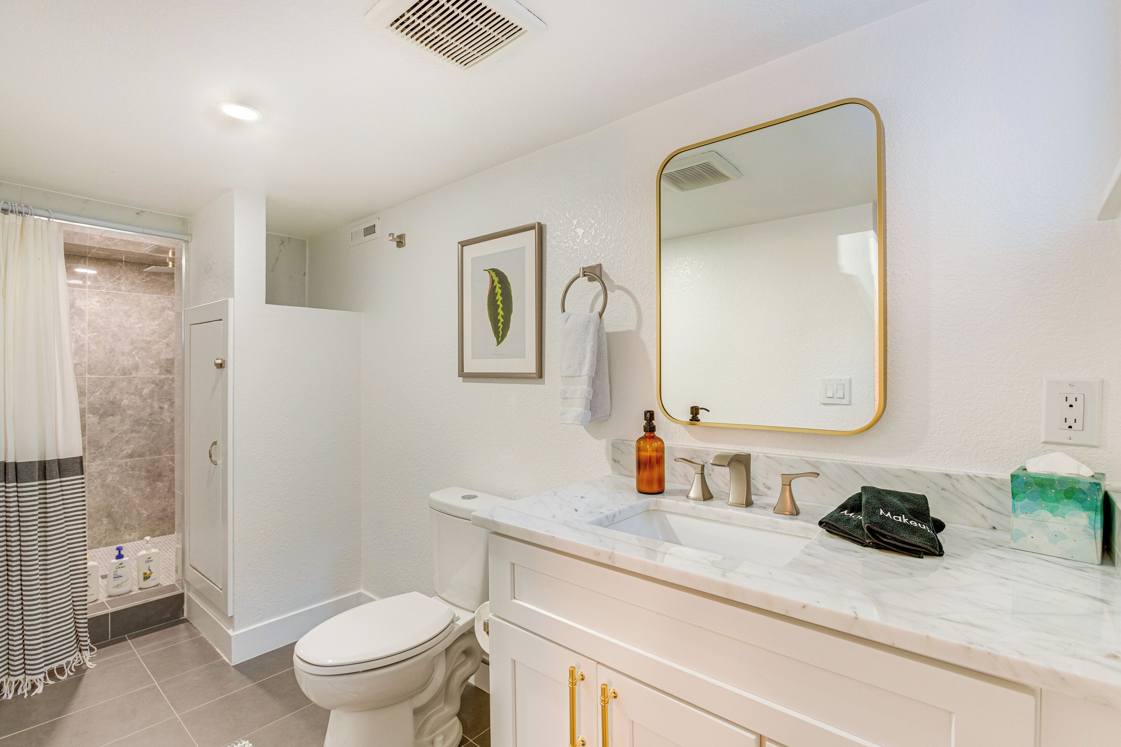 A bathroom with a white toilet, a shower, a marble sink, and a mirror with a gold frame.