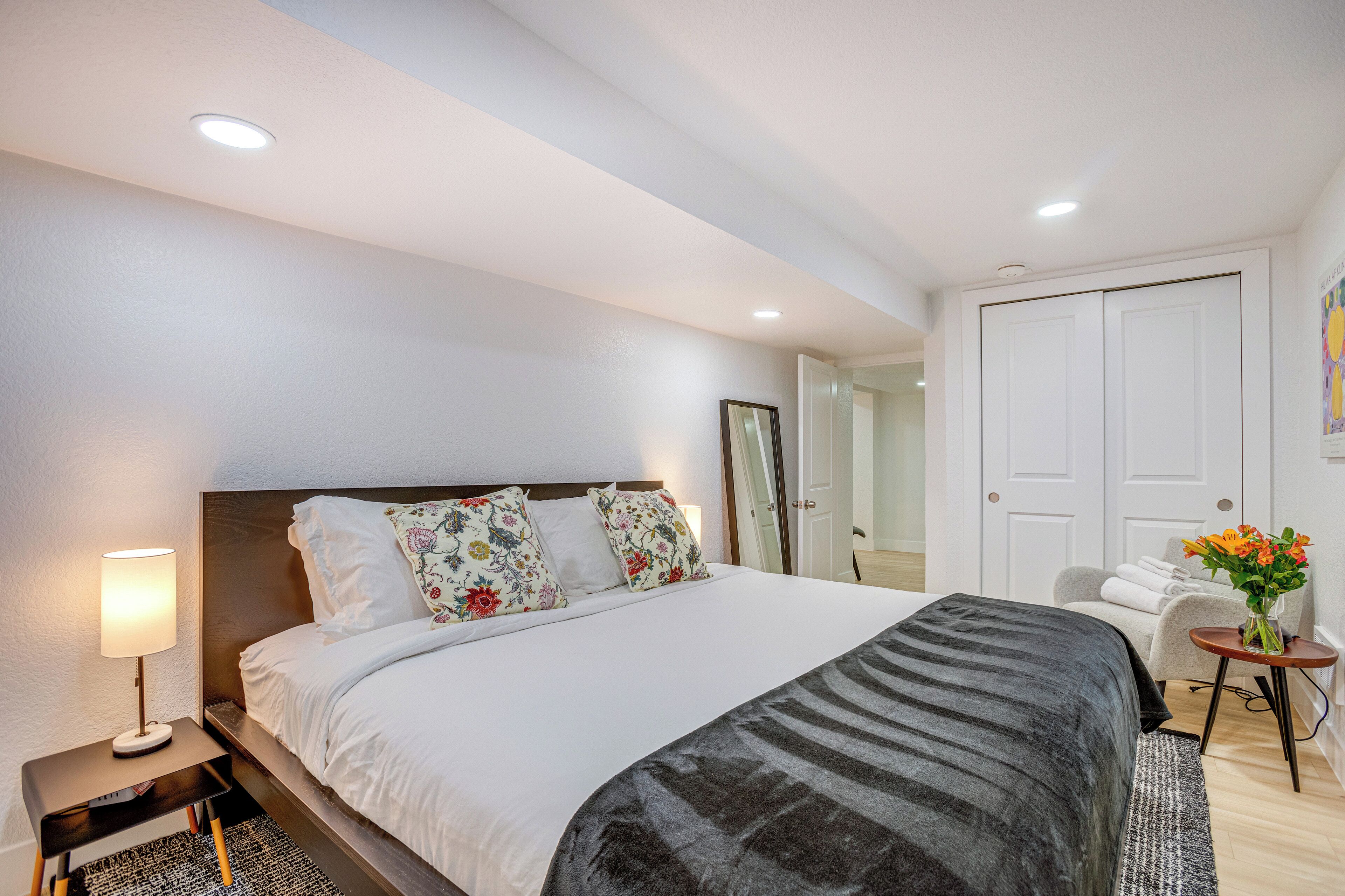 Une chambre d’hôtel moderne avec un grand lit, une table de chevet avec une lampe, une petite table d’appoint avec un vase de fleurs et une porte donnant sur une autre pièce.
