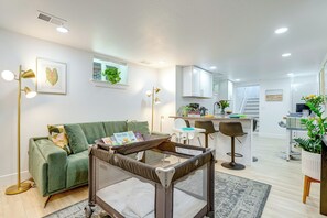 A modern living room with a green sofa, a baby playpen, a dining area with bar stools, and a kitchen in the background. - Wash Park Access: Denver Apt w/ Outdoor Dining! (Denver)