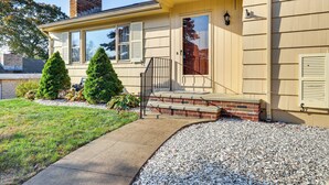 A two-story house with a front porch, a brick chimney, and a small front yard with a gravel path and two evergreen trees.