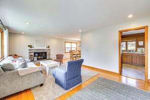 Ein Wohnzimmer mit einer Couch, einem Sessel, einem Couchtisch und einem Kamin. Es gibt eine Essecke mit Stühlen und einem Tisch. Der Raum hat Holzböden und einen Blick zur Küche durch eine offene Tür.