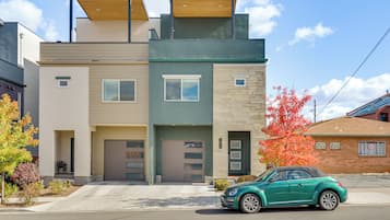 A modern two-story residential building with a garage, a green car parked in front, and a tree with red leaves to the right.