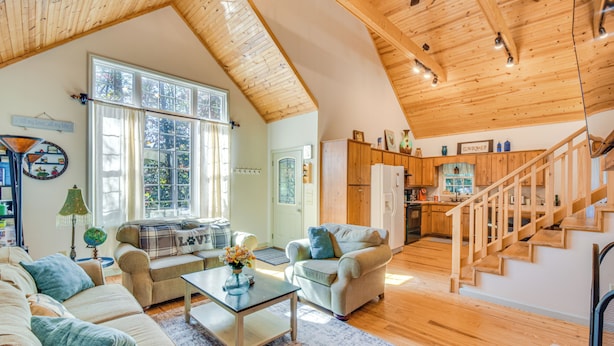Un ampio soggiorno con soffitto alto, travi in legno e una grande finestra. È presente un angolo cottura con mobili in legno e una scala che conduce a un altro livello.