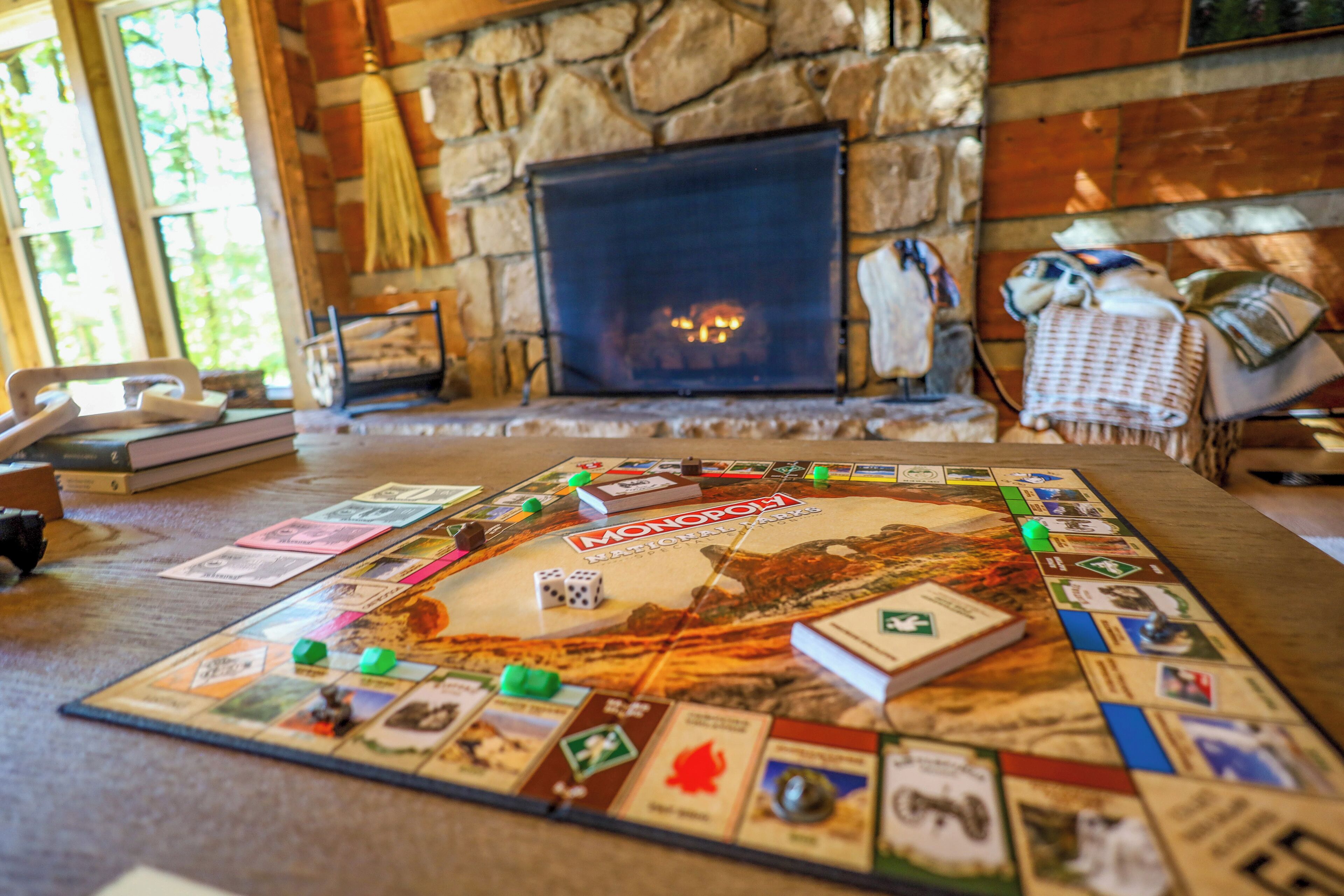 Un jeu de société Monopoly est installé sur une table en bois dans une pièce dotée d’une cheminée en pierre et d’une fenêtre.