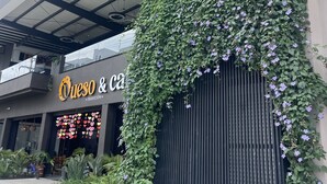 Un bâtiment avec une enseigne indiquant « Queso & ca » et un espace d’entrée abrité, agrémenté d’un mur recouvert de plantes grimpantes vertes.