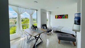 Une chambre moderne comprenant une table à manger, des chaises, un canapé et un lit. De grandes fenêtres sont agrémentées de rideaux, et on y trouve également un distributeur d’eau ainsi qu’une peinture abstraite aux couleurs vives accrochée au mur.