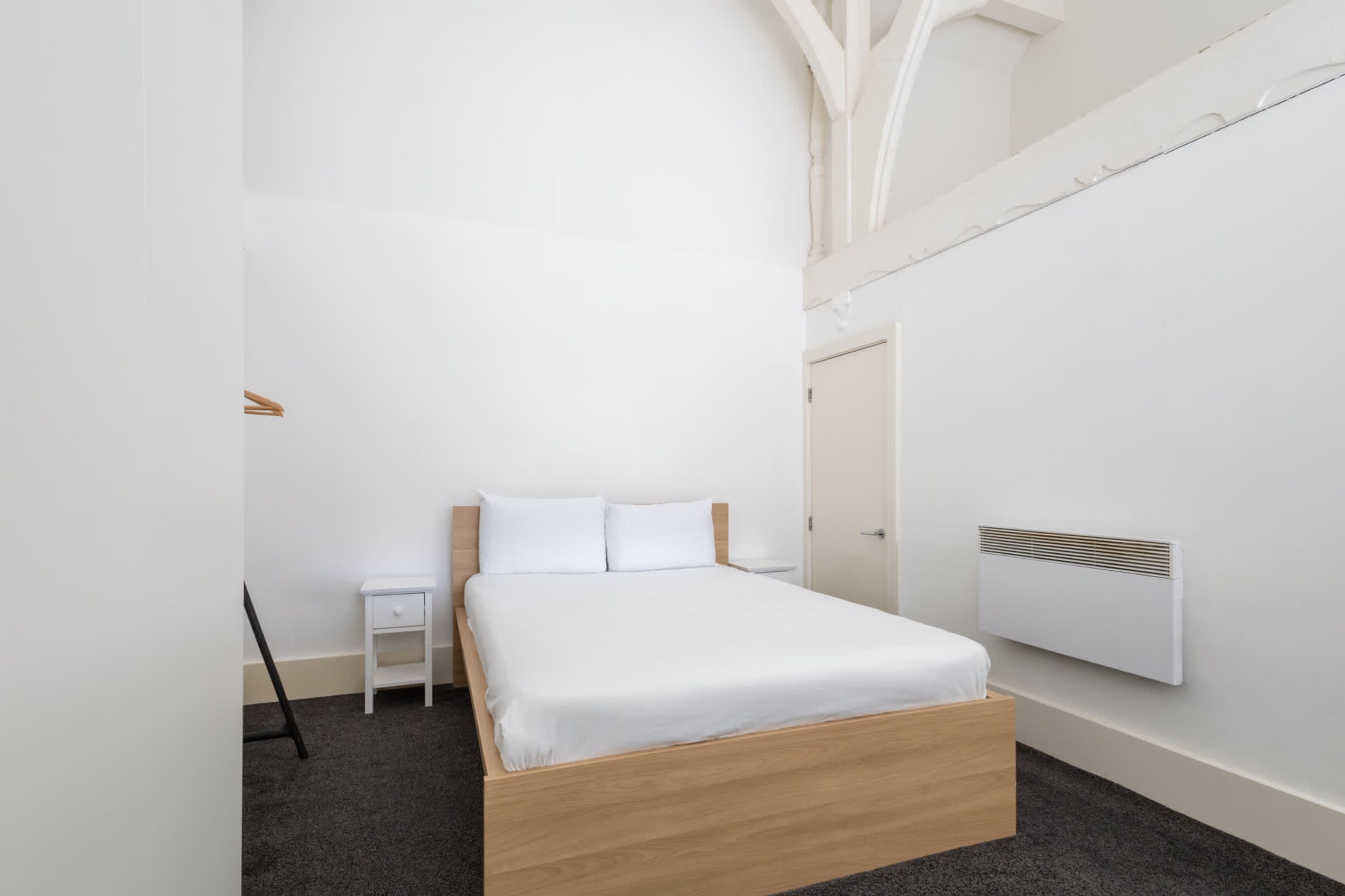 Quarto minimalista com cama de madeira, roupa de cama branca, mesa de cabeceira pequena e aquecedor fixado na parede.