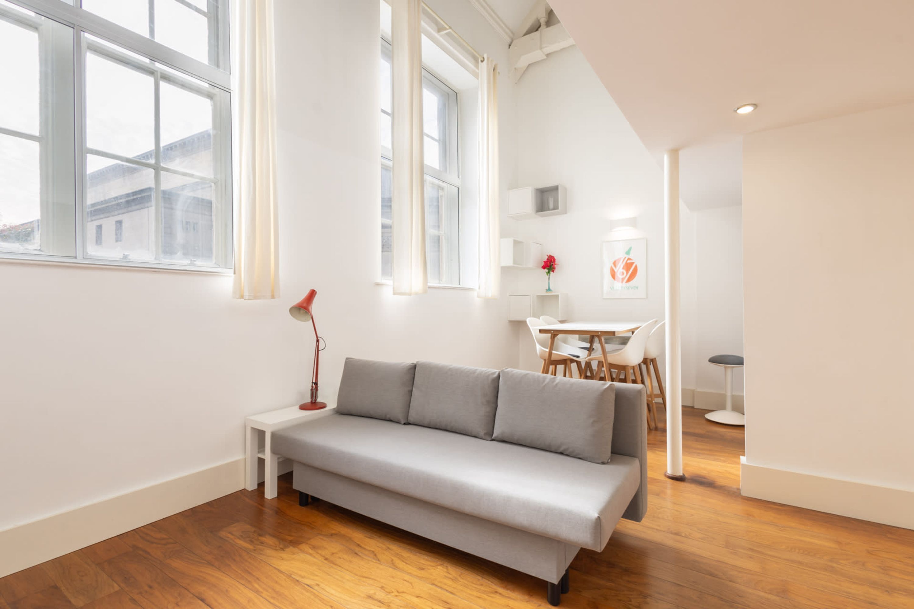 Una sala de estar moderna con un sofá gris, suelo de madera, ventanas amplias y un comedor al fondo.