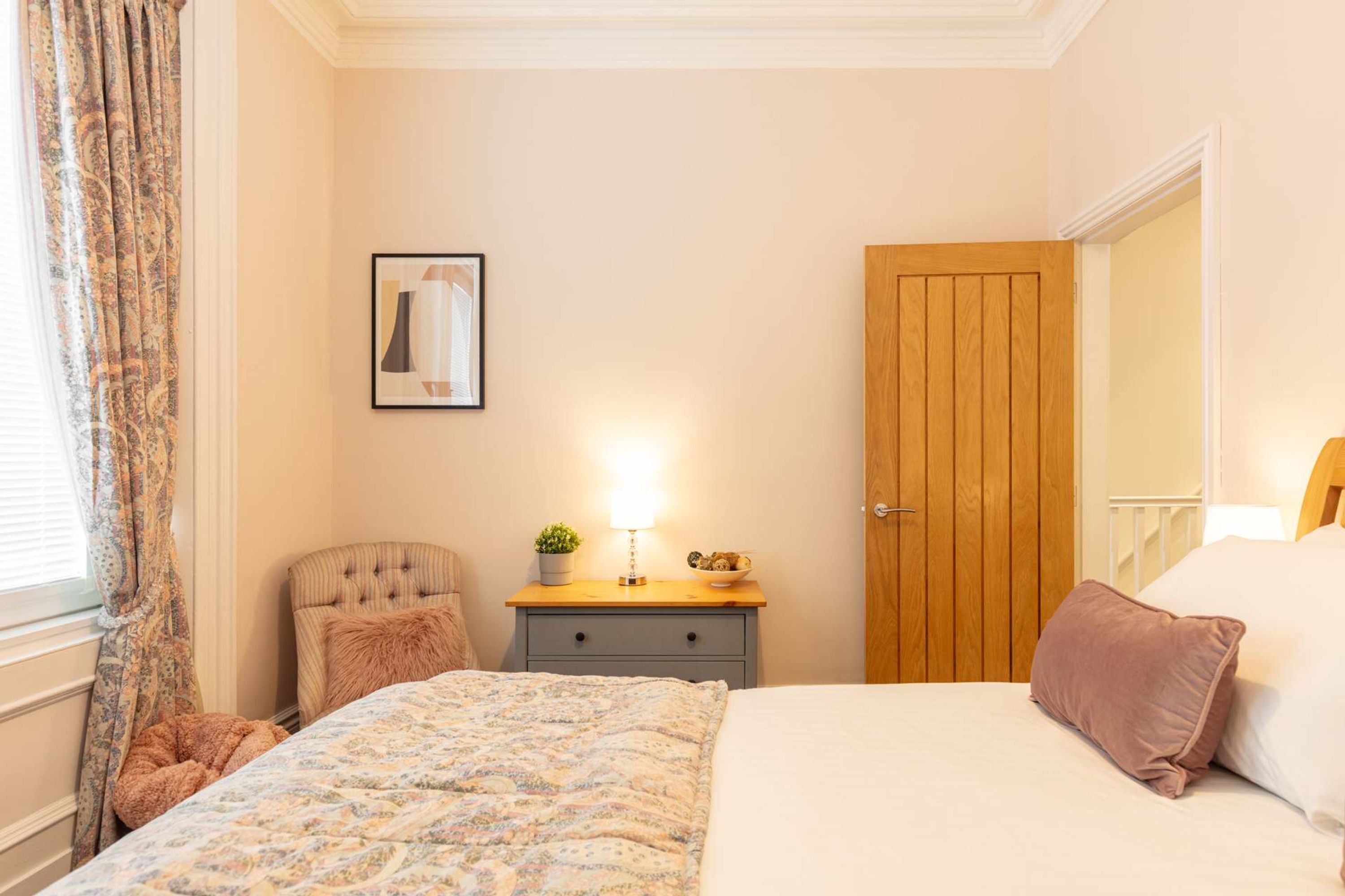 A bedroom with a bed, a chair, a nightstand with a lamp, a wooden door, and a framed picture on the wall.