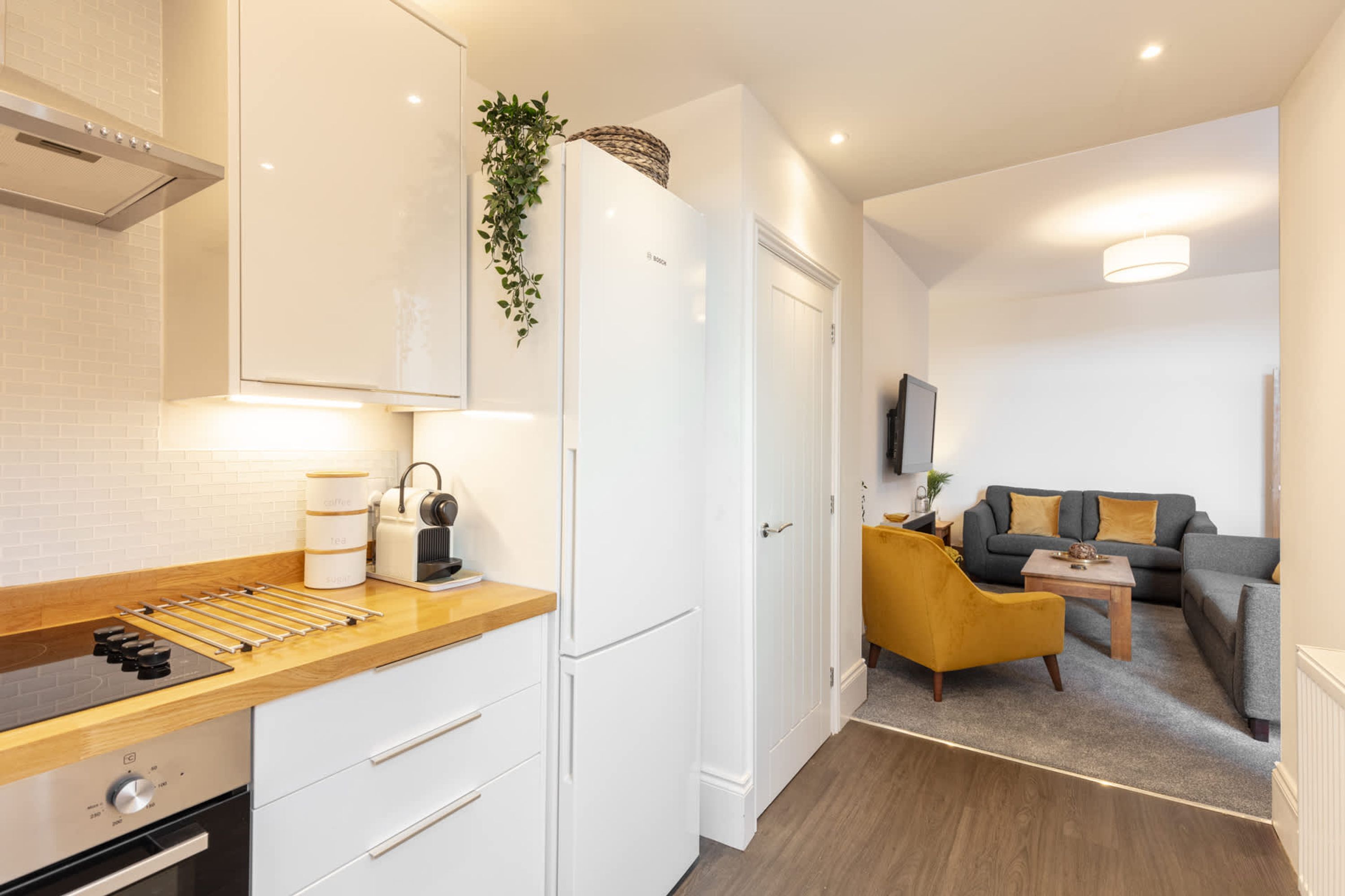 A modern kitchen with white cabinets, a built-in oven, and a wooden countertop. There is a small dining area with a yellow chair and a table.
