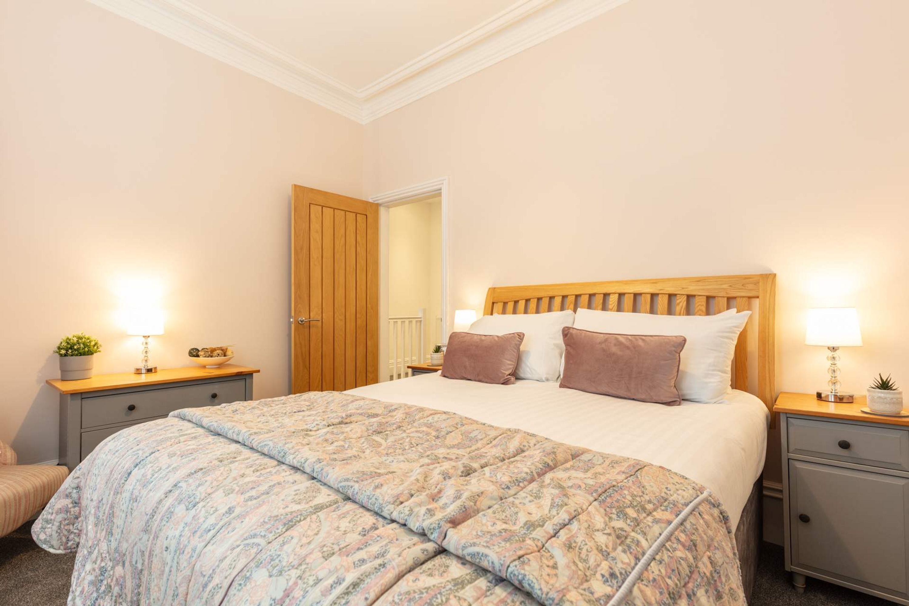Une chambre à coucher avec un grand lit, deux tables de chevet, une porte en bois et une petite plante posée sur l’une des tables.