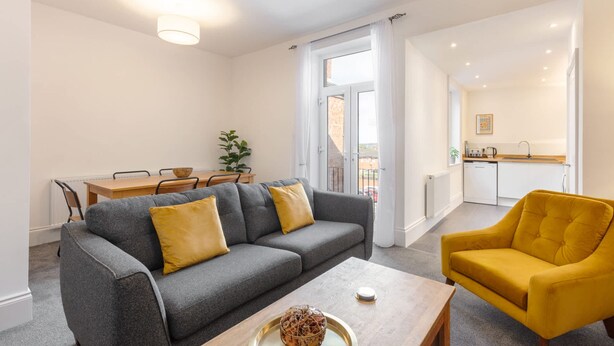 Un salon moderne avec un canapé gris, des coussins jaunes, une table basse en bois et un fauteuil jaune. On y trouve également un coin repas avec une table et des chaises en bois, et une cuisine à l’arrière-plan.