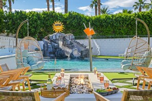A poolside area with a rock waterfall, lounge chairs, a fire pit, and a table set for dining. - WOW The Hamptons Pickleball Massive Pool Slide (Indio)