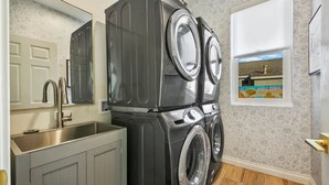 Pequeña lavandería con lavadora y secadora apiladas, un lavamanos y una ventana que da a una casa.