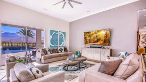 A spacious living room with a large window, a ceiling fan, and a flat-screen TV mounted on the wall. - The Royal Bahamian- Pickleball Golf Sleeps 20 (Indio)