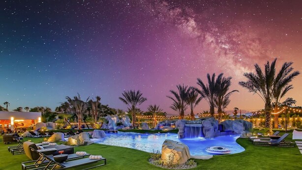 A nighttime resort pool area with a clear view of the Milky Way, surrounded by palm trees and lounge chairs.