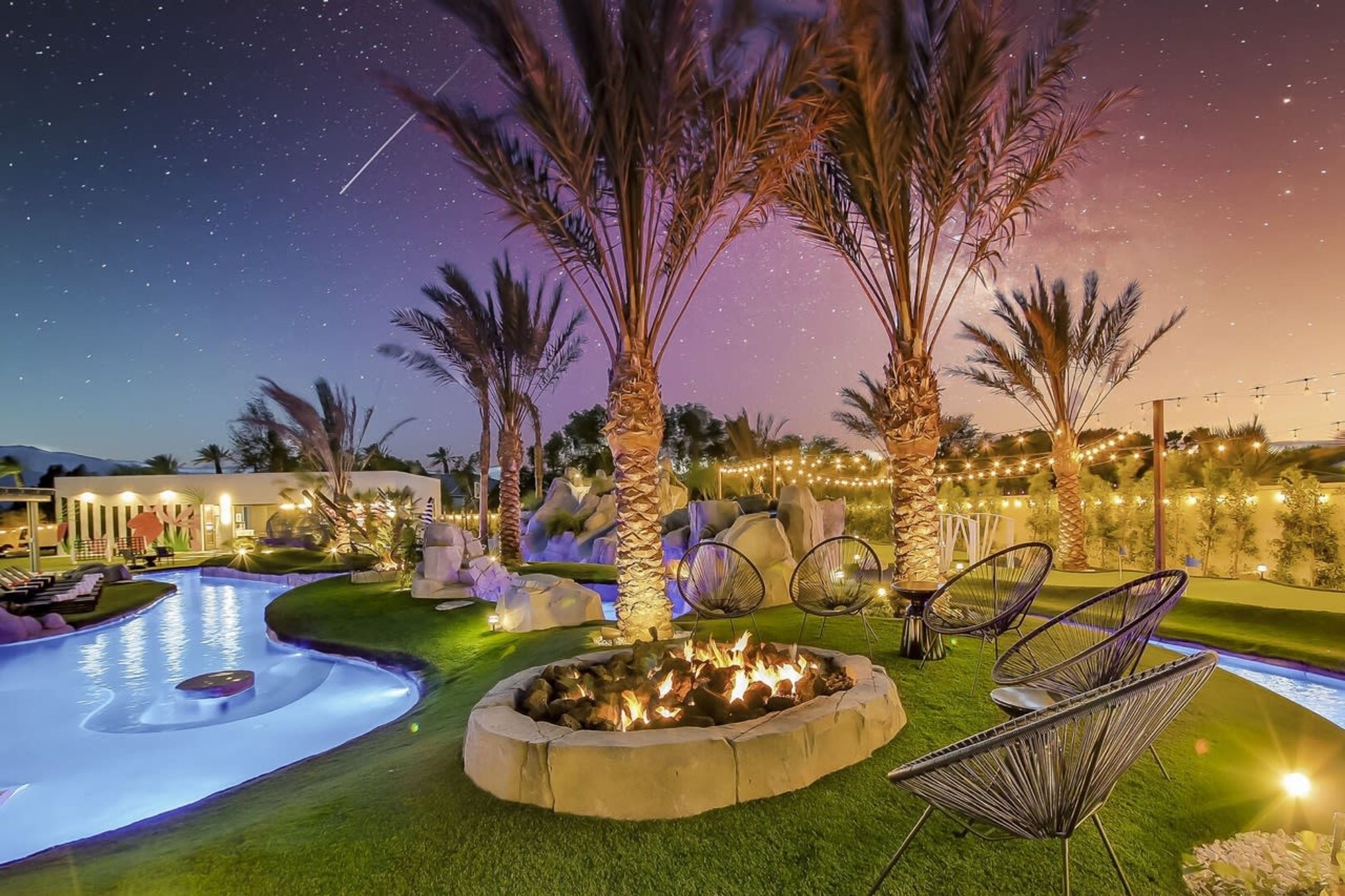 A nighttime outdoor pool area with a fire pit, lounge chairs, and string lights.