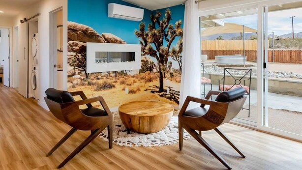 A modern living room with a desert mural, two leather chairs, a wooden coffee table, and a large window offering a view of a fenced outdoor area.