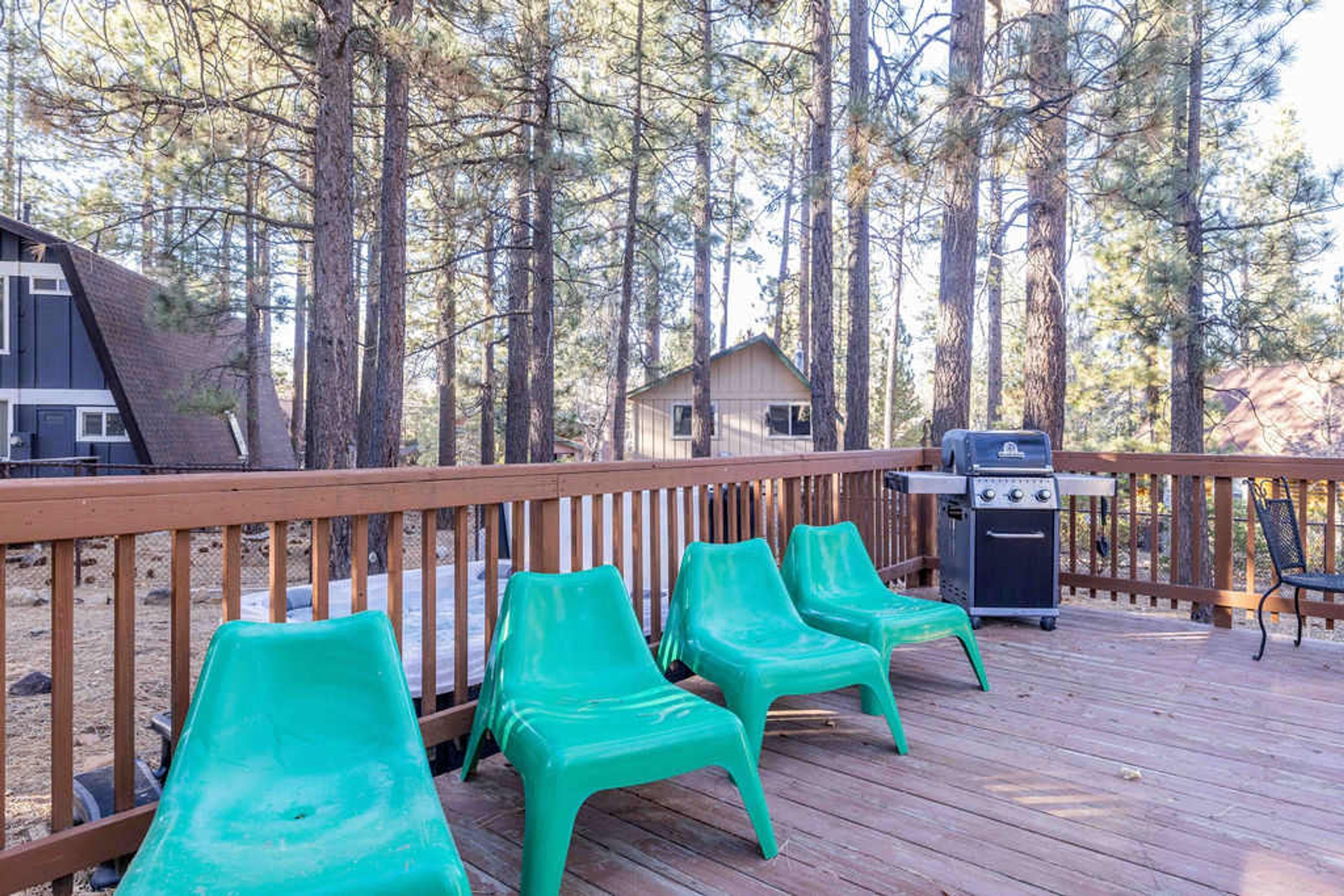 A wooden deck with green plastic chairs, a grill, and a small cabin in a forested area.