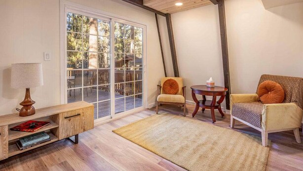 Un salón acogedor con suelo de madera, un amplio ventanal corredero que da a un espacio exterior, una mesita de madera, dos sillas y una mesita auxiliar con una lámpara.