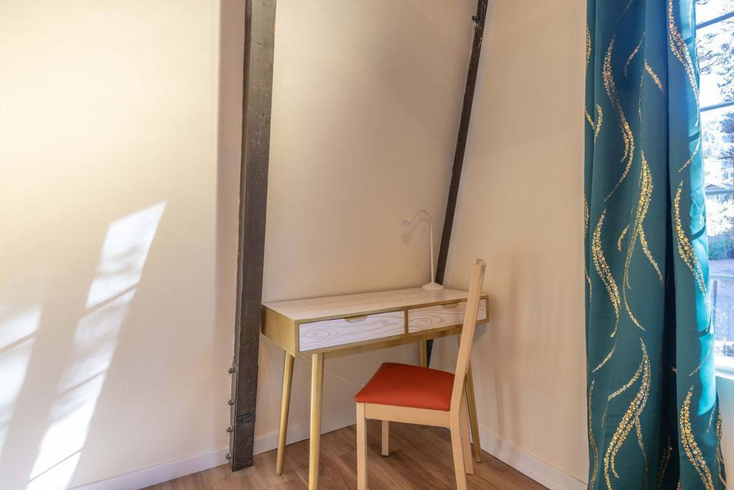 Een hoek kamer met een bureau en stoel, een raam met gordijnen, en een houten vloer.
