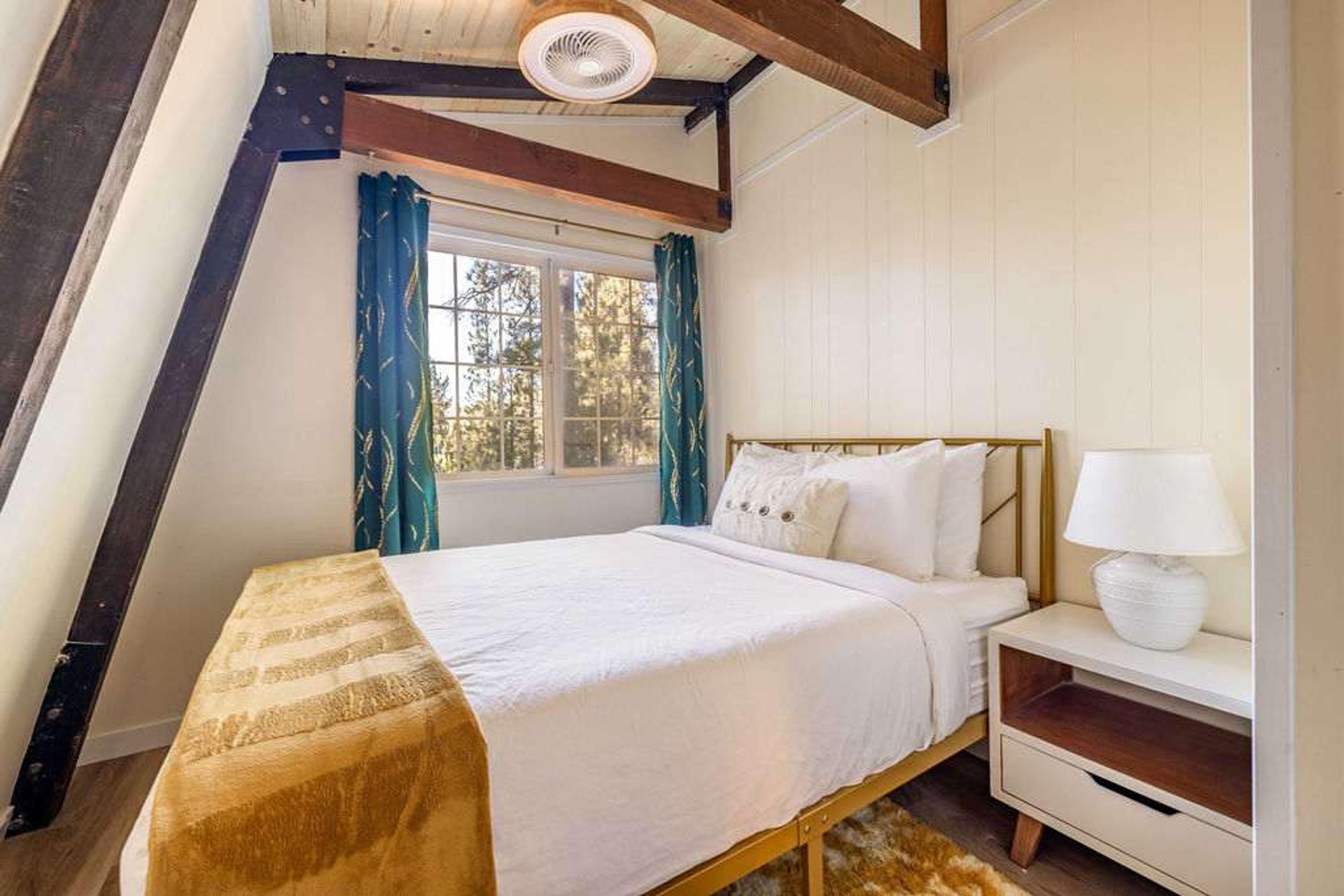 Ein Schlafzimmer mit Bett, Nachttisch, Lampe, einem Fenster mit Vorhängen und einem Deckenventilator.