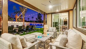 An outdoor patio area with seating, a television displaying a golf course, and string lights. - Forever Young Crocslide Pickleball 7BD Game Rm (Indio)