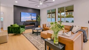 A modern living room with a fireplace, green and brown sofas, a coffee table, and a large TV screen displaying a lighthouse. - Forever Young Crocslide Pickleball 7BD Game Rm (Indio)