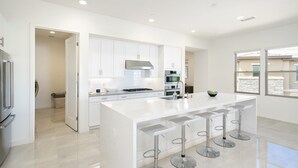 Una cocina moderna con armarios blancos, una isla central con taburetes de bar, electrodomésticos de acero inoxidable y ventanas amplias que dejan entrar luz natural.