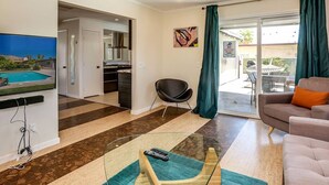 Una sala de estar moderna con un televisor de pantalla plana, una mesa de centro de vidrio y un sofá. Se ve un área de cocina a través de una puerta abierta, y un balcón con vistas.