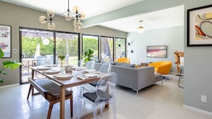 A modern living room with a grey sofa, a dining table set with clear chairs, and a large window offering a view of the outdoors. - Lux Desert Retreat 4BR Pool Spa Stunning Views (Palm Springs)