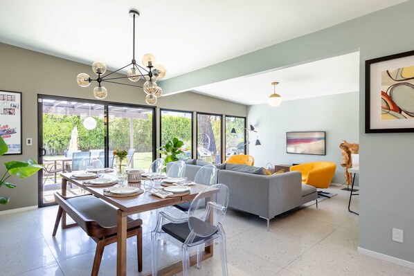 A modern living room with a grey sofa, a dining table set with clear chairs, and a large window offering a view of the outdoors.